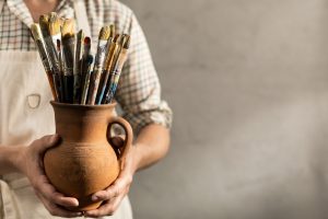 Male artist holding clay jug with paintbrush. Man artist painter and paint brush in creative studio